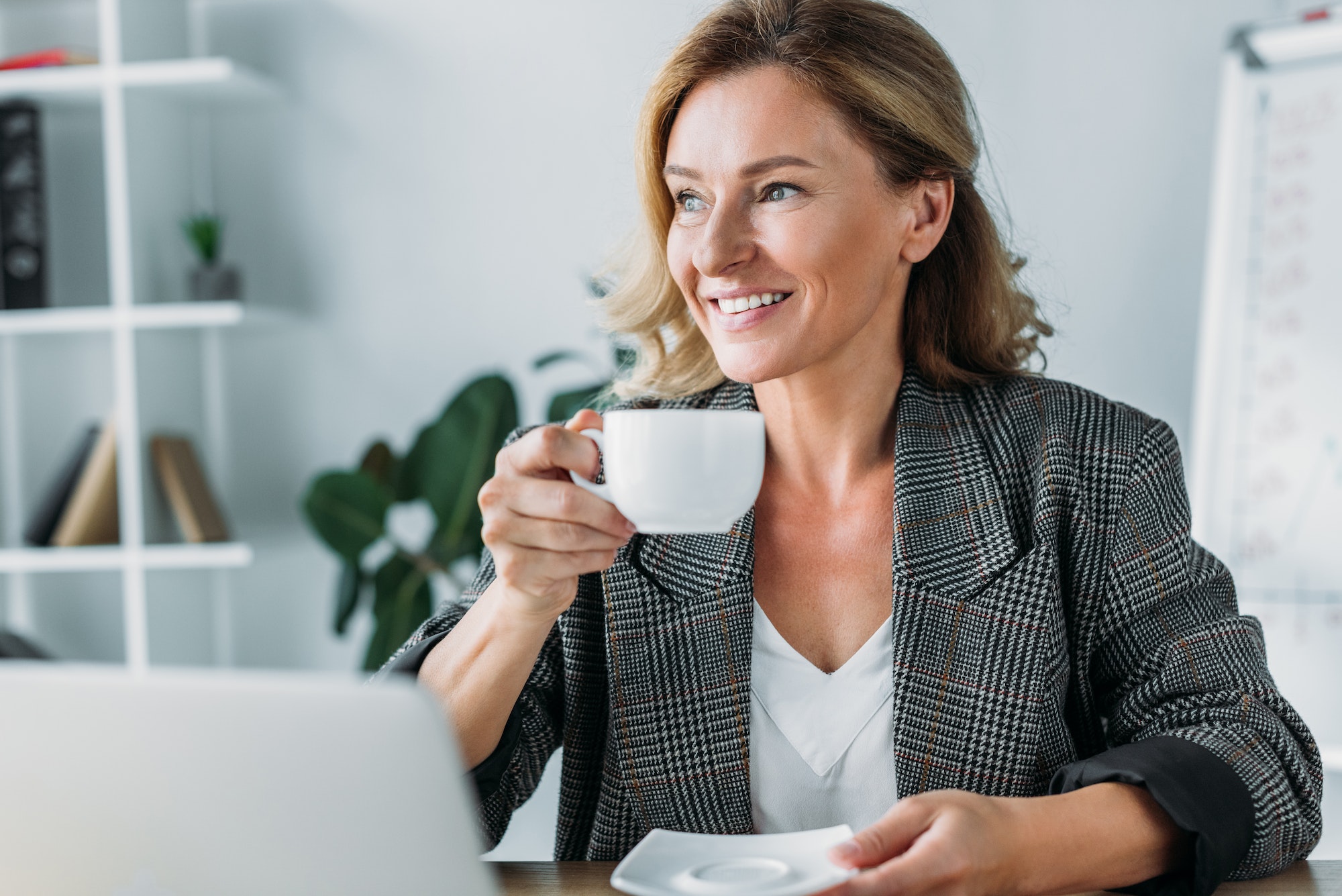 aantrekkelijke onderneemster zittend met kop koffie aan tafel in kantoor en wegkijkend